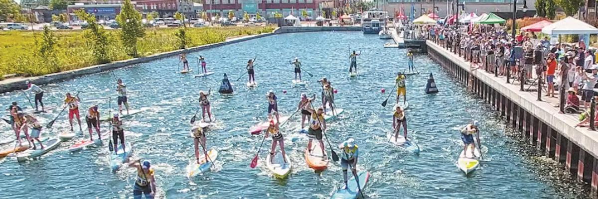 Group paddleboarding event on a sunny day in a canal, with participants paddling while crowds watch from the nearby boardwalk.