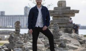 Jon Montgomery, host of The Amazing Race Canada stands in front of an Inukshuk with Collingwood Grain Terminals in the distance