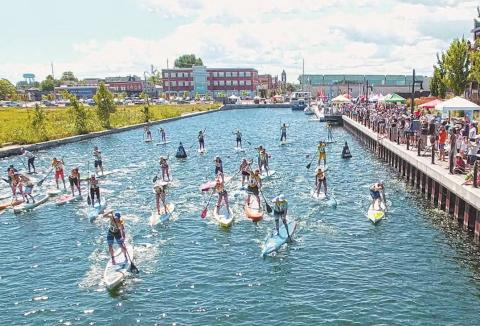 Group paddleboarding event on a sunny day in a canal, with participants paddling while crowds watch from the nearby boardwalk.